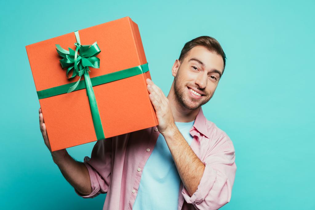 Gülümseyen adam büyük bir hediye kutusu tutuyor, mavi üzerine izole edilmiş. - Fotoğraf, Görsel