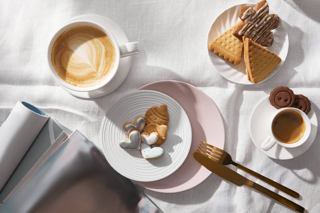 Kaffeetassen von oben neben Keksen und Zeitschriften auf weißer Tischdecke - Foto, Bild
