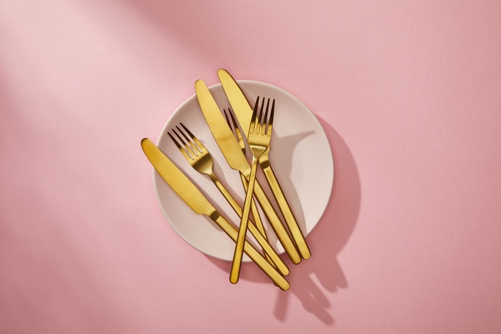 Bovenaanzicht van glanzend goudkleurig bestek op lege plaat op roze achtergrond - Foto, afbeelding