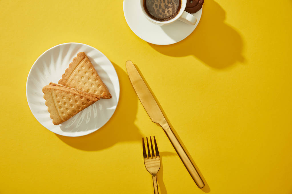Κάτοψη του φλιτζανιού του καφέ με χρυσά μαχαιροπίρουνα και νόστιμα μπισκότα στην κίτρινη επιφάνεια - Φωτογραφία, εικόνα