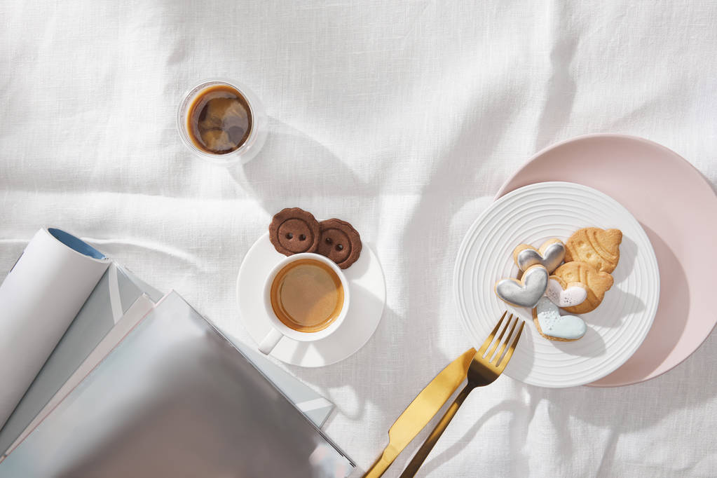 Κάτοψη φρέσκου καφέ, μπισκότο με γλάσο και περιοδικά σε λευκό τραπεζομάντηλο - Φωτογραφία, εικόνα
