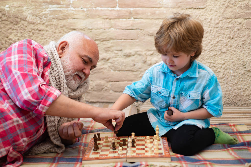 Ο παππούς και ο εγγονός παίζουν σκάκι και χαμογελούν ενώ περνούν χρόνο μαζί στο σπίτι. Ώριμος άντρας με μικρό αγόρι να παίζει σκάκι.. - Φωτογραφία, εικόνα