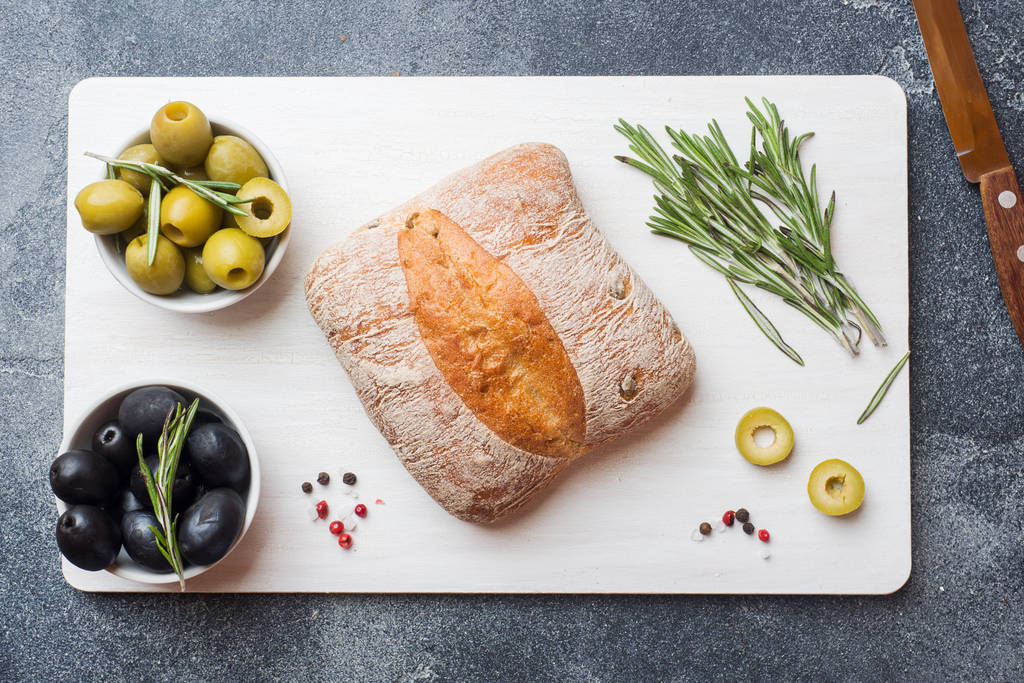 italienisches Ciabatta-Brot mit Oliven und Rosmarin auf einem Schneidebrett. dunkler Betonhintergrund. - Foto, Bild