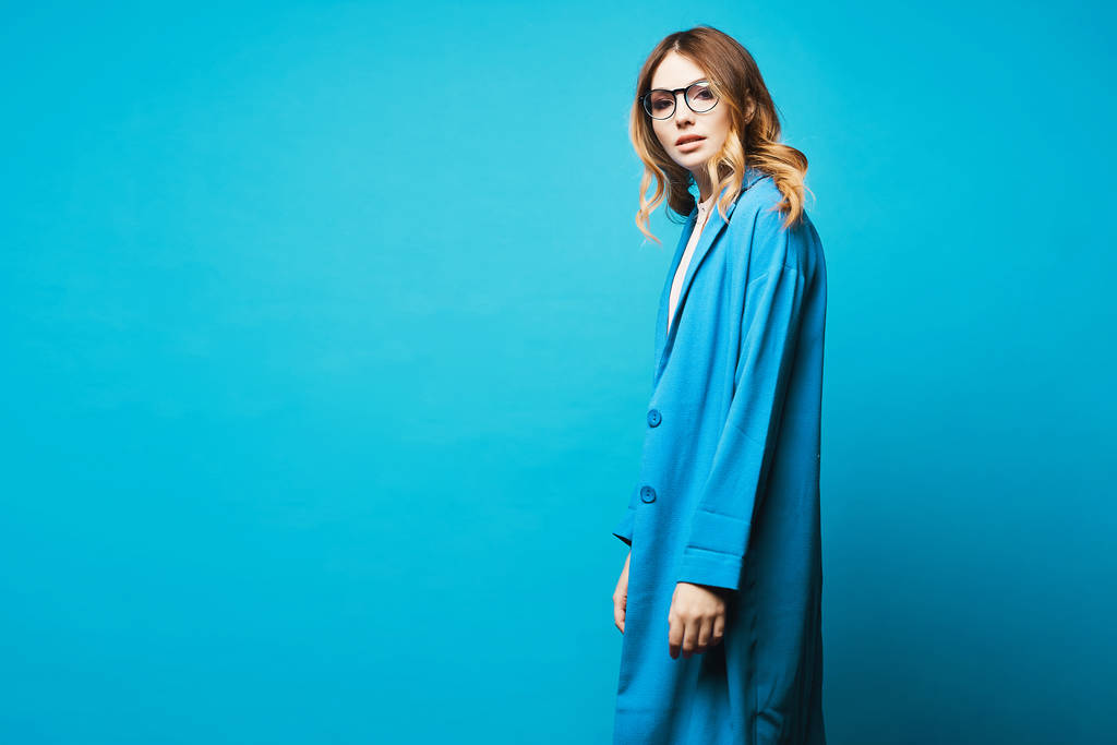 Beautiful young woman with natural makeup in stylish glasses and trendy blue coat, isolated. Stylish model girl in blue coat and modish eyewear at blue background, isolated. Copy space. Female fashion - Photo, Image
