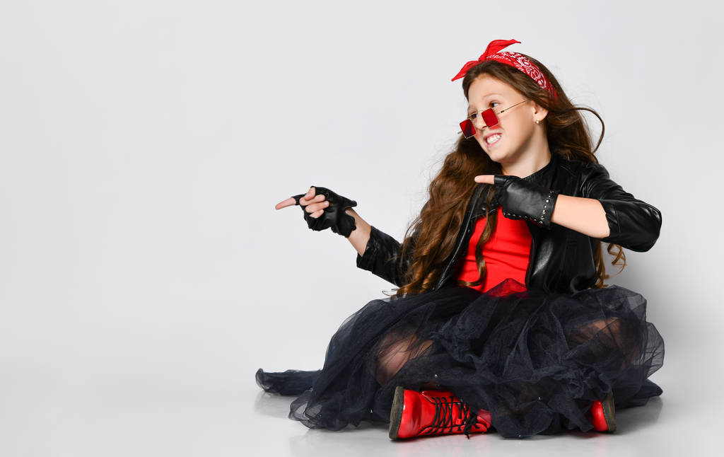 junges positives Mädchen Modell in heller Rock-Style lässiger Kleidung und roten Stiefeln auf dem Boden sitzend und mit den Fingern zur Seite zeigend - Foto, Bild
