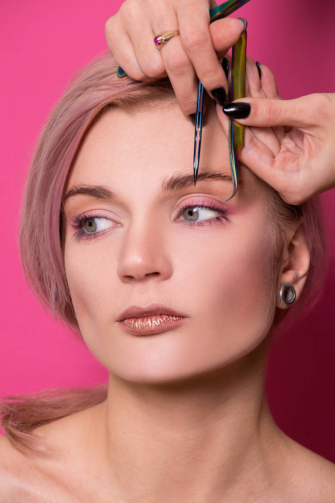 Primer plano de una hermosa chica de apariencia europea sostiene en su mano pinzas para las cejas cerca de la cara en el estudio aislado sobre un fondo rosa
 - Foto, imagen