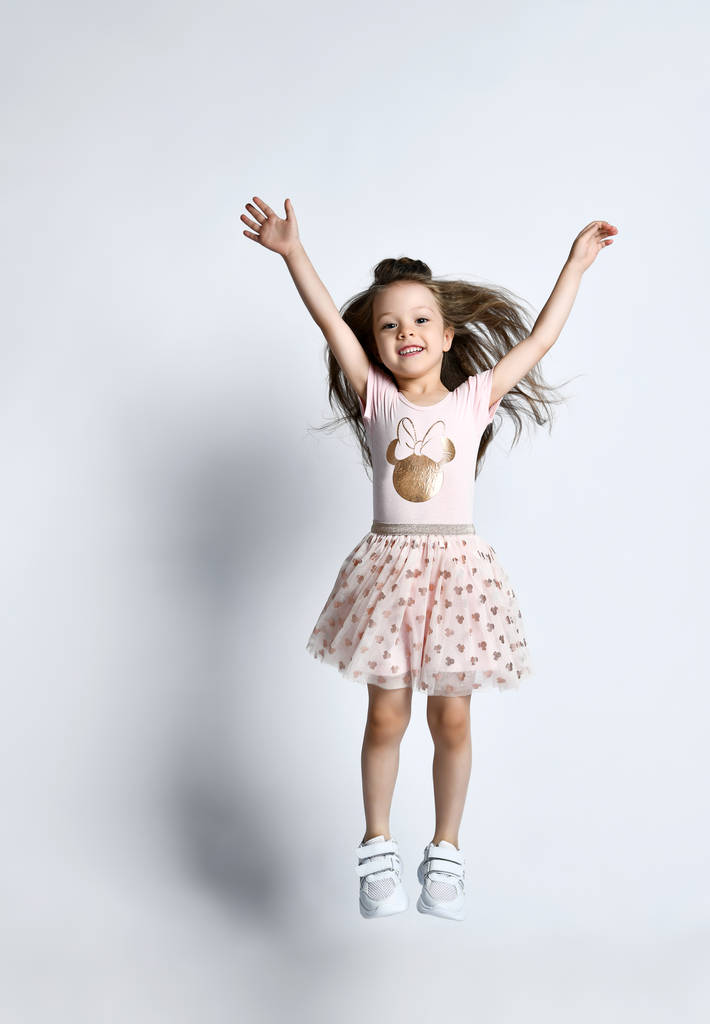 kleines lächelndes nettes Mädchen in lässigem Sommerkleid und Turnschuhen, die über weißen Wandhintergrund springen - Foto, Bild