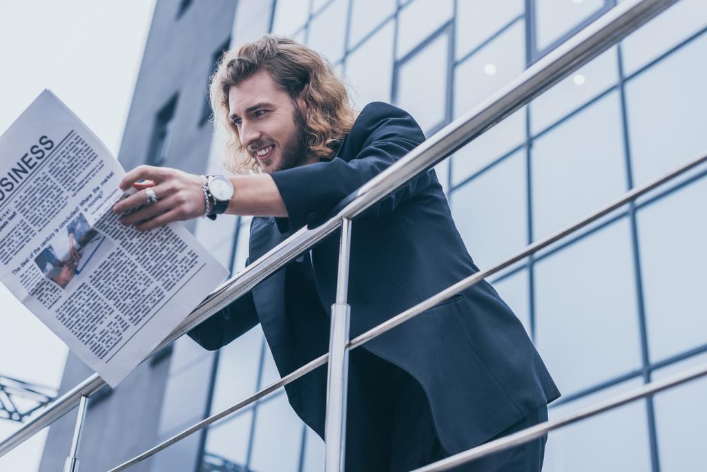 Низкий угол зрения улыбающийся модный бизнесмен в черном костюме читает деловую газету возле офисного здания и перила
 - Фото, изображение