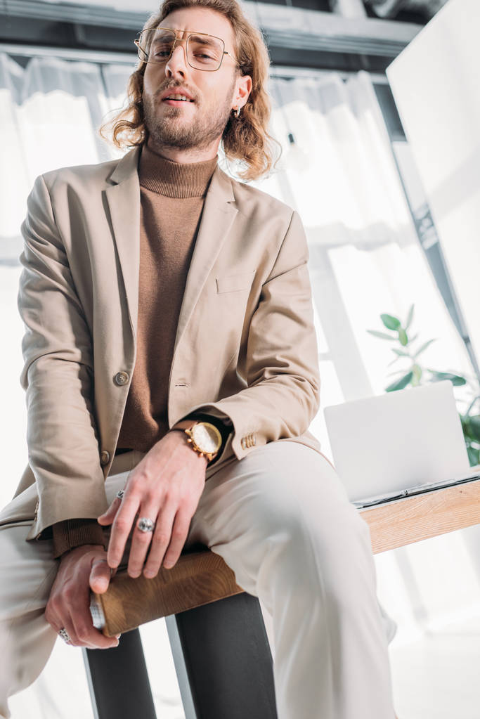 низький кут зору впевнений модний бізнесмен сидить на столі і позує в офісі
 - Фото, зображення