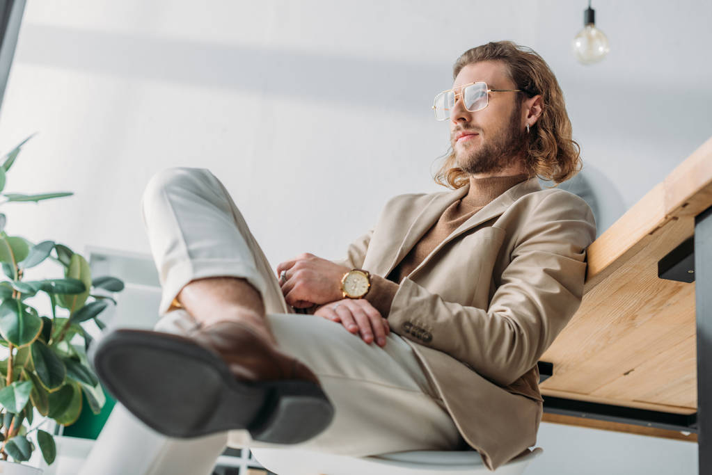 χαμηλή γωνία άποψη της κομψής μόδας επιχειρηματίας κάθεται στην καρέκλα κοντά στο γραφείο στο γραφείο - Φωτογραφία, εικόνα