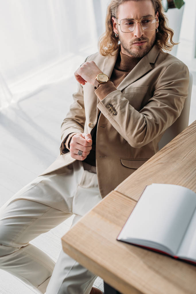 selective focus of pensive fashionable businessman sitting on chair near desk with blank notebook in office - Photo, Image