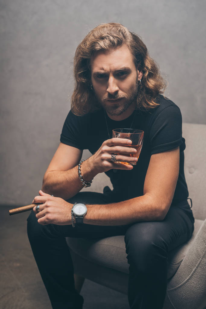 μοντέρνος επιχειρηματίας με μαύρα ρούχα καπνίζει πούρο και πίνει ουίσκι σε πολυθρόνα κοντά σε γκρι τοίχο - Φωτογραφία, εικόνα