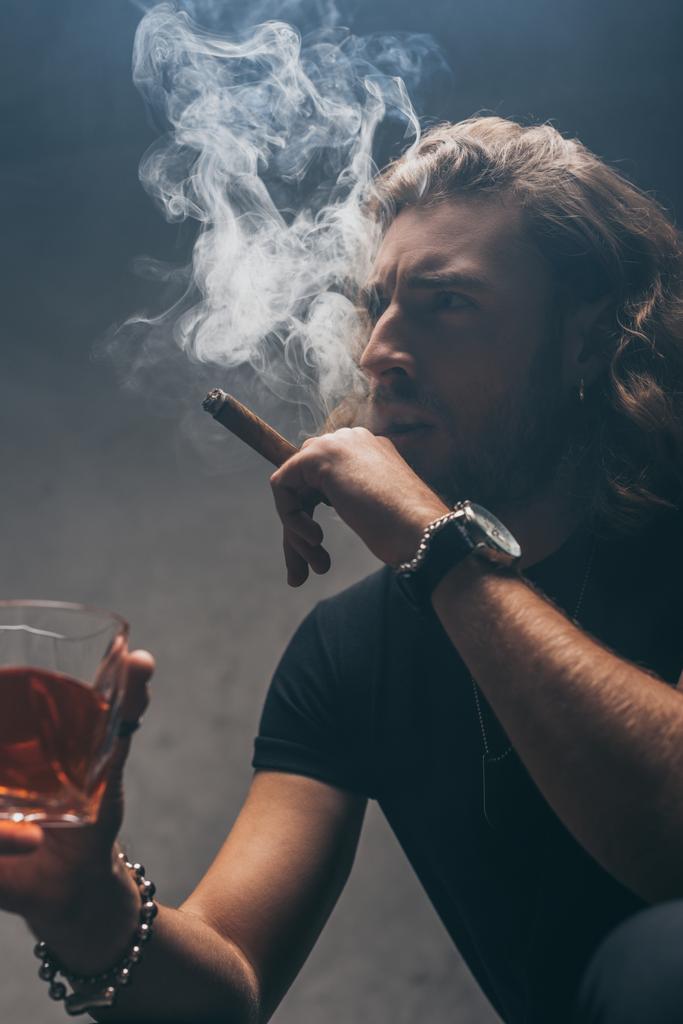 jännittynyt muodikas liikemies mustassa asussa tupakointi sikari ja juo viskiä harmaan seinän lähellä
 - Valokuva, kuva