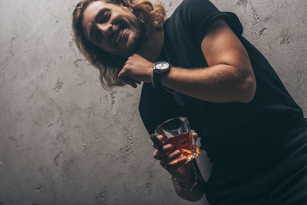 low angle view of smiling fashionable businessman in black outfit with cigar and whiskey near grey wall - Photo, Image