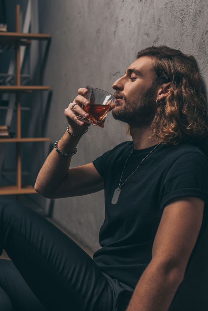 side view of fashionable businessman in black outfit drinking whiskey on floor near grey wall - Photo, Image