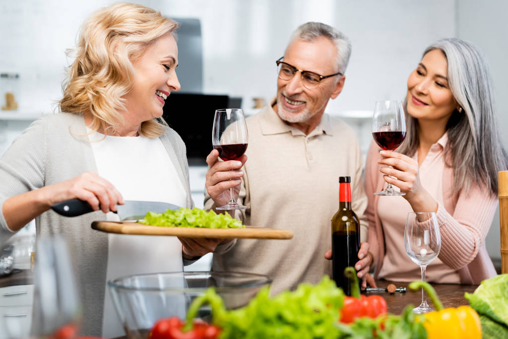 smiling woman holding cutting board with lettuce and her multicultural friends holding wine glasses - Photo, Image