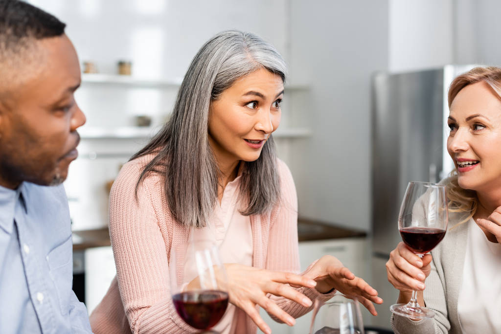 valikoiva painopiste hymyilevä monikulttuurinen ystävät puhuvat ja pitämällä viinilasit keittiössä
  - Valokuva, kuva