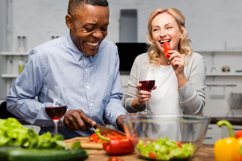 smiling african american man cutting bell pepper and woman holding wine glass and eating bell pepper - Photo, Image