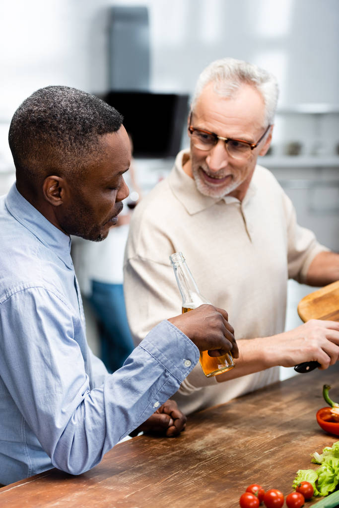 africký Američan drží pivo a jeho usmívající se přítel vaření v kuchyni  - Fotografie, Obrázek