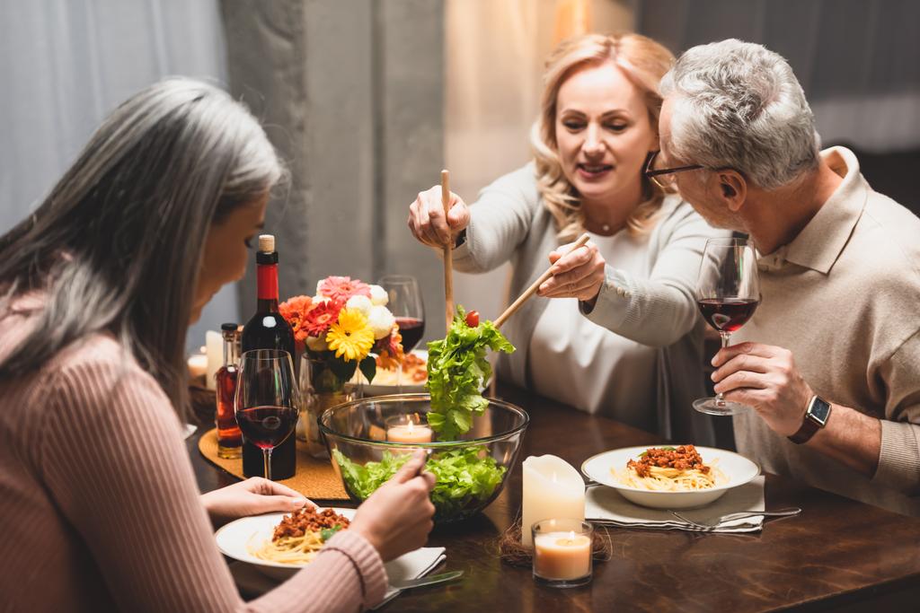 улыбающаяся женщина берет салат из миски и разговаривает с другом во время ужина
  - Фото, изображение