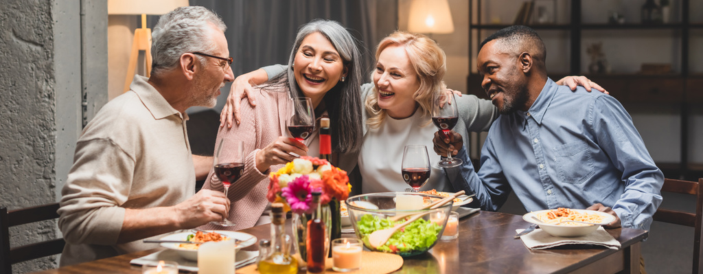夕食中にワイングラスを抱いたり抱き合ったりする多文化の友人たちの笑顔のパノラマ写真  - 写真・画像