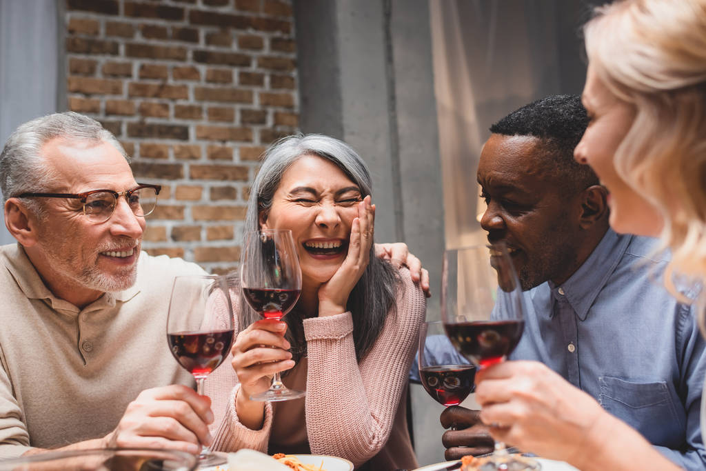 amigos multiculturales sonrientes abrazando y sosteniendo copas de vino durante la cena
  - Foto, Imagen