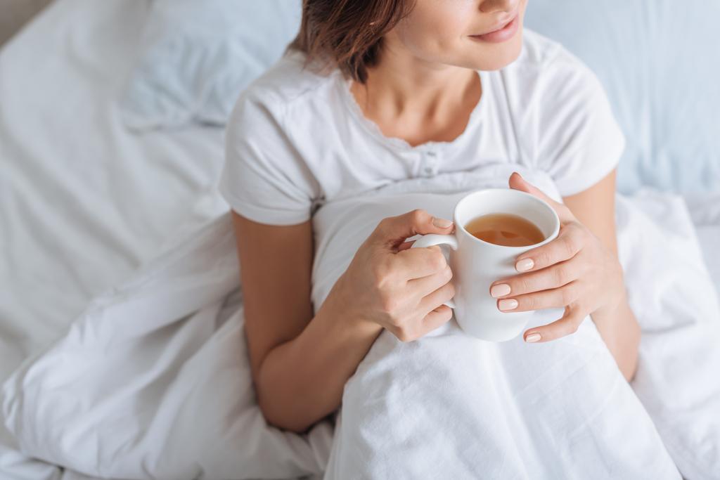 καλλιεργημένη άποψη του ονειρική γυναίκα κρατώντας κύπελλο με τσάι στο κρεβάτι  - Φωτογραφία, εικόνα
