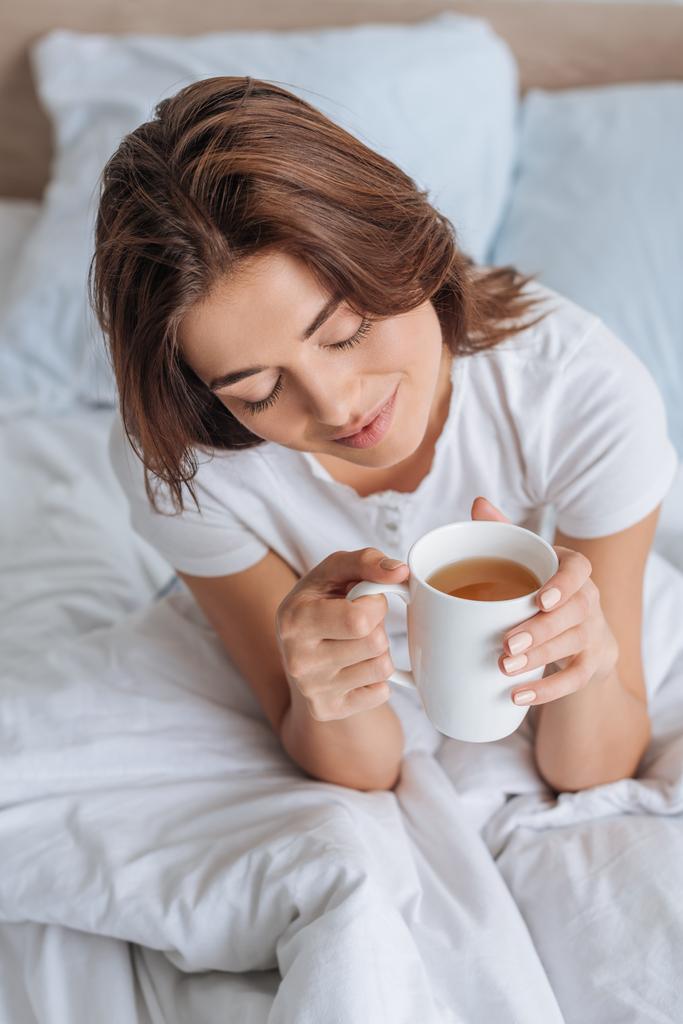 overhead view of happy girl with with closed eyes holding cup with tea in bed  - Photo, Image