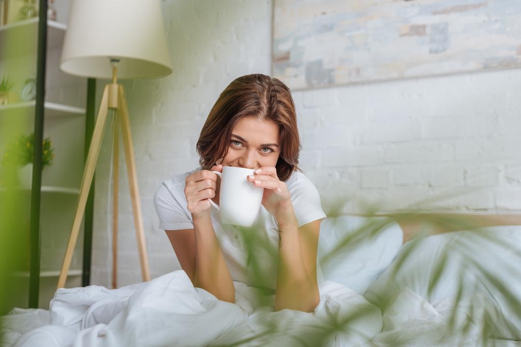 вибірковий фокус молодої жінки, дивлячись на камеру, тримаючи чашку з чаєм у ліжку
  - Фото, зображення