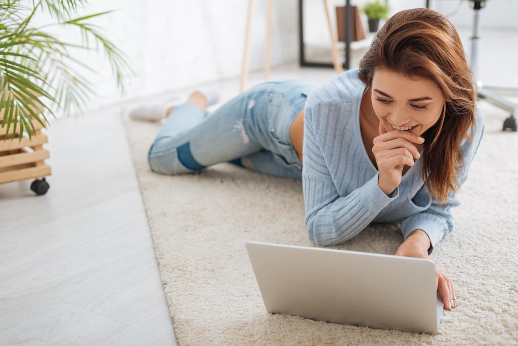 cheerful girl using laptop and smiling while lying on carpet at home  - Photo, Image