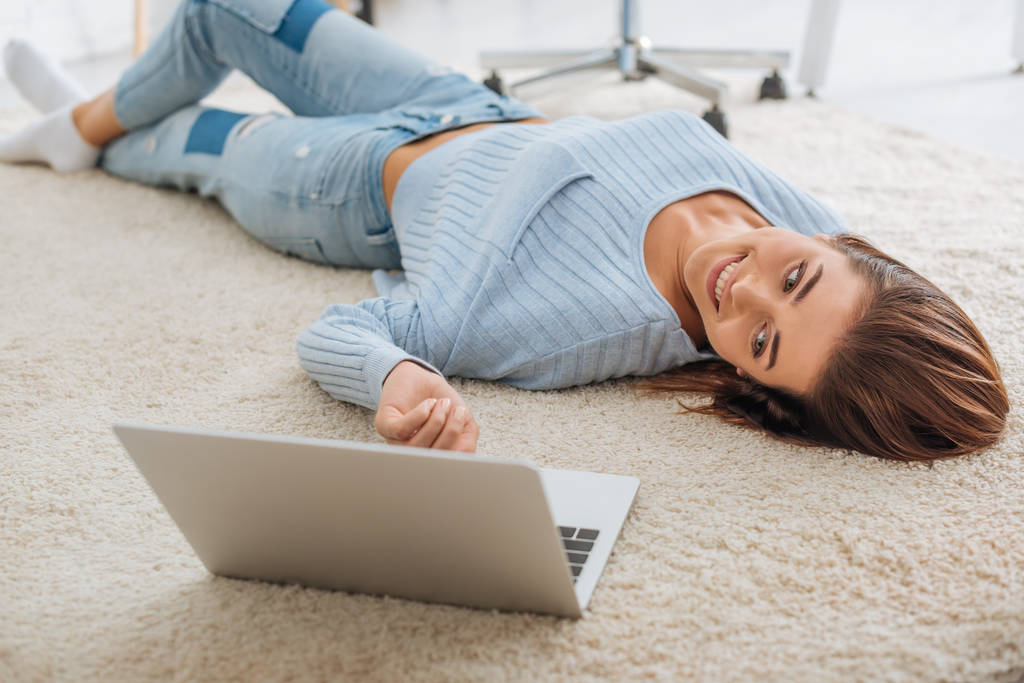 selective focus of cheerful girl smiling while lying on carpet near laptop  - Photo, Image