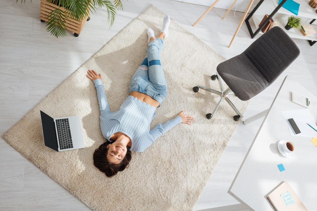 górny widok pozytywnej kobiety odpoczywającej podczas leżenia na dywanie w pobliżu laptopa w domu  - Zdjęcie, obraz