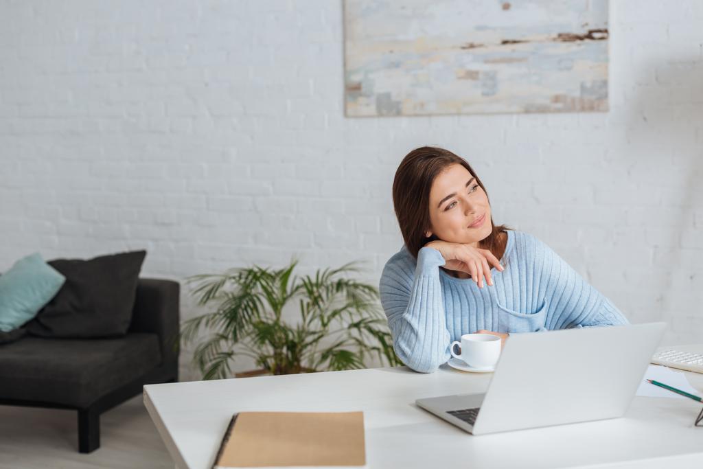 dreamy woman looking away near laptop and cup on table  - Photo, Image