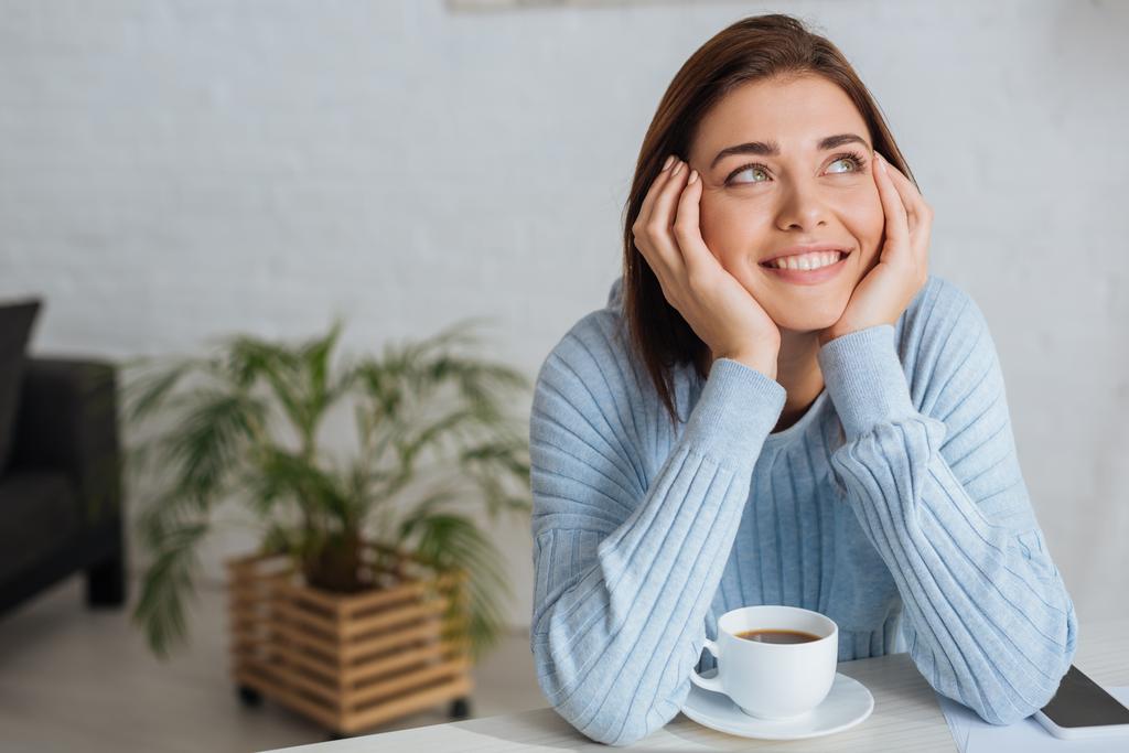 ονειρική νεαρή γυναίκα χαμογελά κοντά κύπελλο με καφέ  - Φωτογραφία, εικόνα