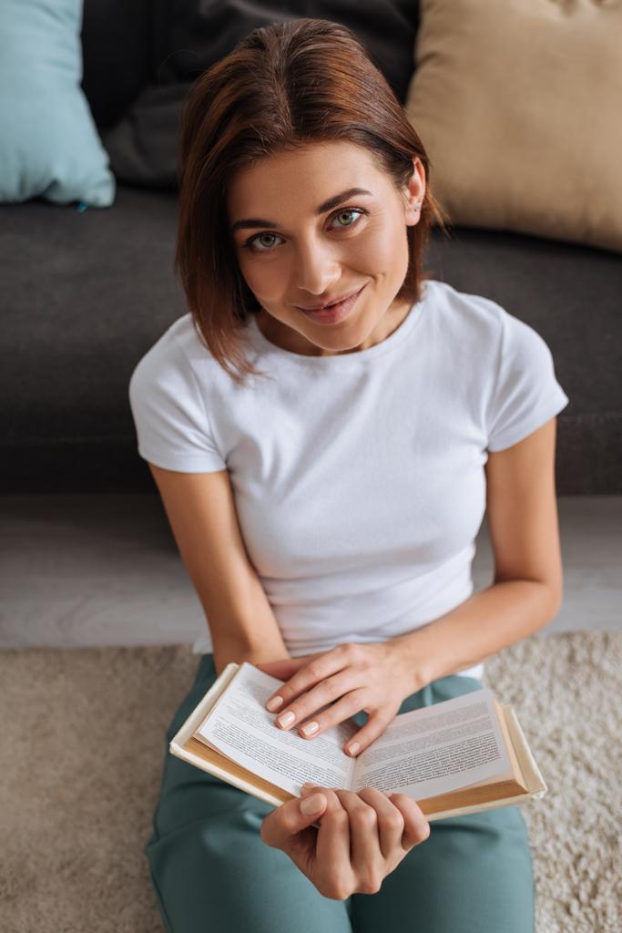 happy woman holding book while sitting on carpet near sofa  - Photo, Image