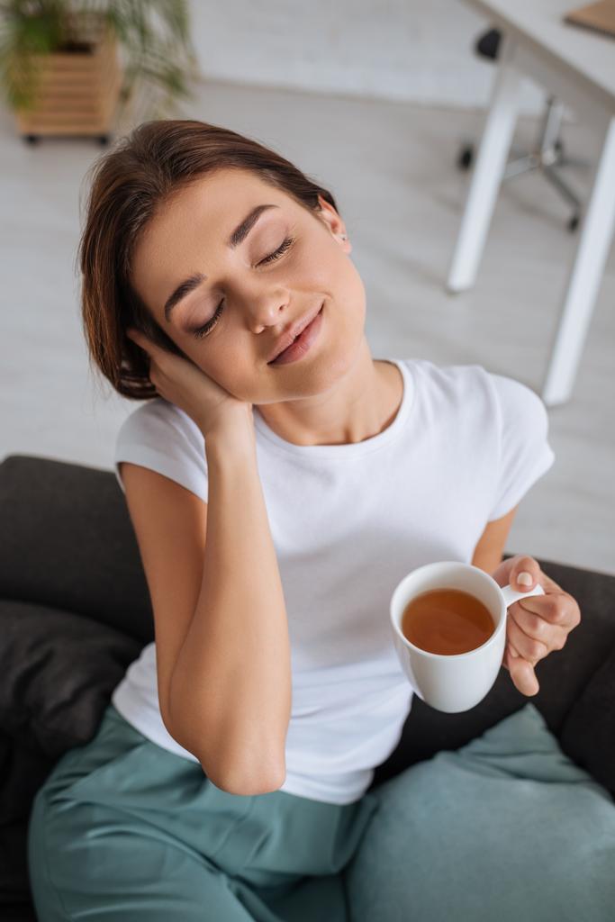 junge verträumte Frau hält Tasse mit Tee, während sie auf dem Sofa im Wohnzimmer chillt  - Foto, Bild
