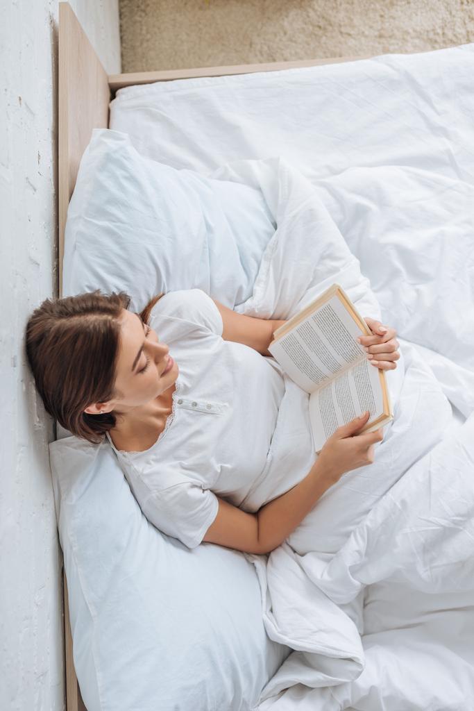 top view of happy girl reading book while chilling in bed  - Photo, Image