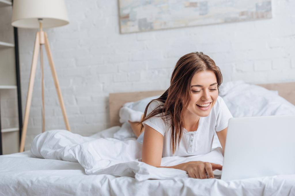 gelukkig jong vrouw met behulp van laptop terwijl chillen in slaapkamer  - Foto, afbeelding