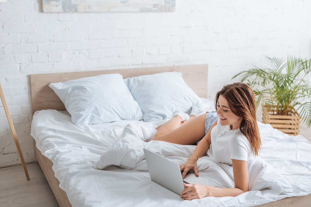 alegre joven mujer usando el ordenador portátil mientras se enfría en el dormitorio
  - Foto, Imagen