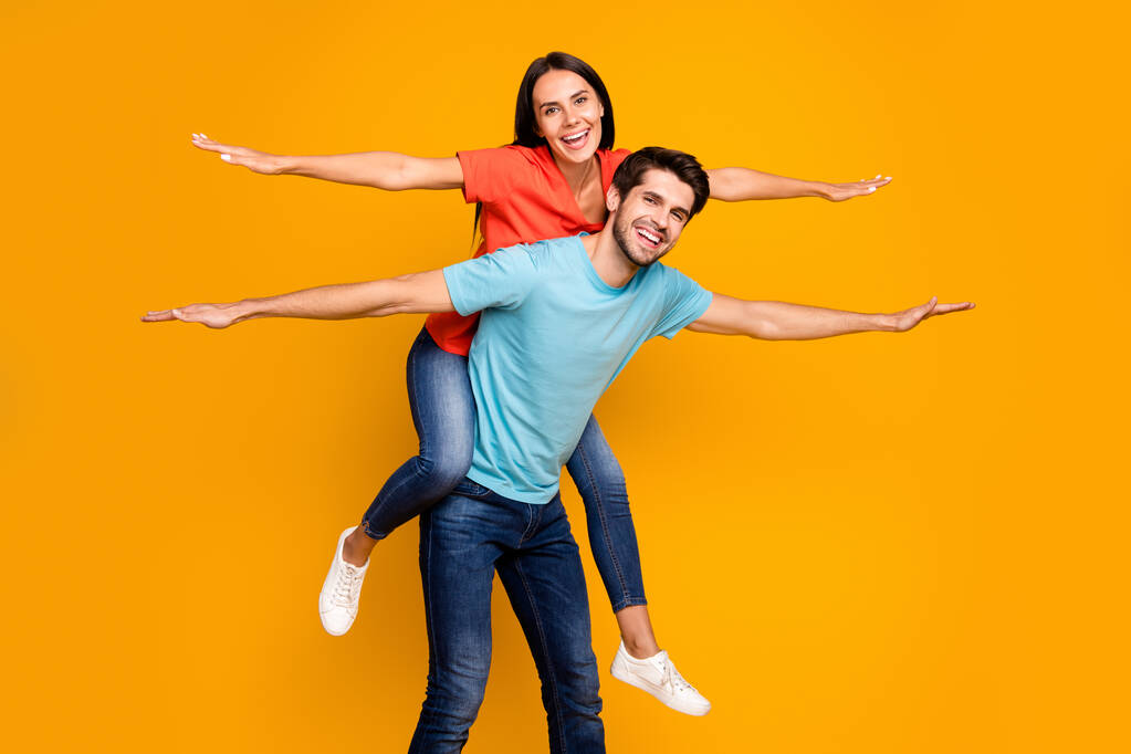 Photo of two funny people guy carry lady piggyback meet summer time together spread hands like wings wear casual stylish blue orange t-shirts jeans isolated yellow color background - Photo, Image