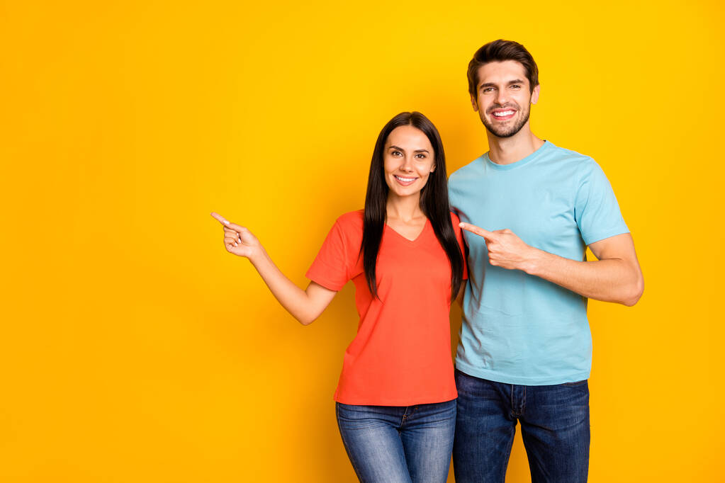 Foto de divertido dos personas pareja chico y señora abrazo dirección dedos vacío espacio precios bajos compras desgaste casual azul naranja camisetas jeans aislados color amarillo fondo
 - Foto, imagen