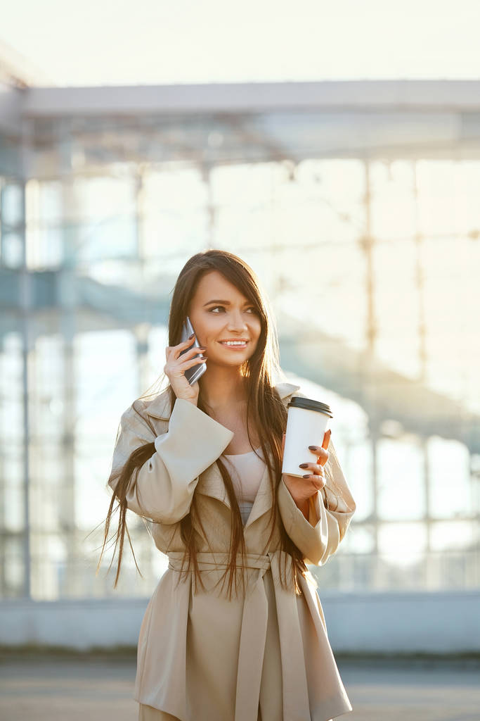  Happy Woman aan de telefoon, staande op straat. Portret van mooie glimlachende vrouwelijke kop koffie in de hand, praten op telefoon buiten. Meisje met smartphone. - Foto, afbeelding