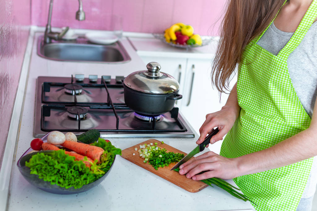 Vrouw in schort bij het fornuis en het snijden van groenten op snijplank voor soep in de keuken. Koken voorbereiding voor het diner. Schoon gezond voedsel en goede voeding.  - Foto, afbeelding