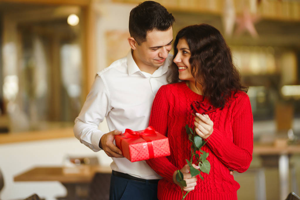 Adam karısına kırmızı kurdeleli bir hediye kutusu veriyor. Sevgi dolu bir çift restoranda sarılıp sevgililer gününü kutluyor. Sevgililer Günü, tatil ve sürpriz konsept. İlişki ve aşk. - Fotoğraf, Görsel