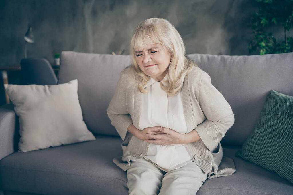 Zdjęcie białowłosej babci w rozpaczy trzymając brzuch cierpiących na zaburzenia hormonalne siedzi sofa divan pokryte koc kraty salon w pomieszczeniach - Zdjęcie, obraz