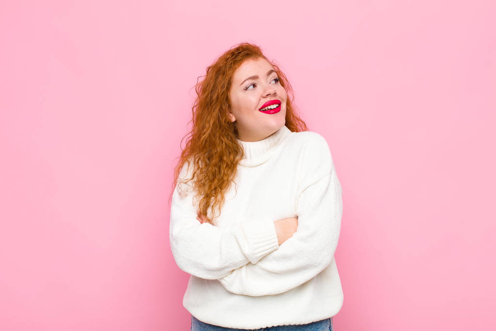 jonge roodharige vrouw die zich gelukkig, trots en hoopvol voelt, zich afvraagt of denkt, opkijkt om ruimte te kopiëren met gekruiste armen tegen een roze muur - Foto, afbeelding