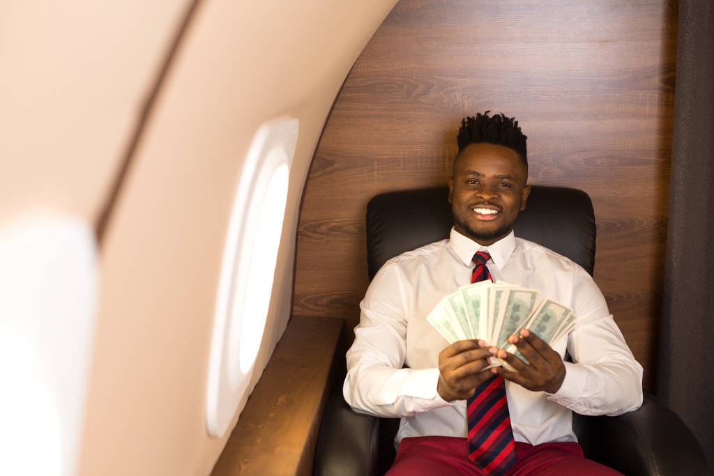 jóképű afrikai férfi fehér ingben egy repülőgép székben, dollárral a kezében. - Fotó, kép
