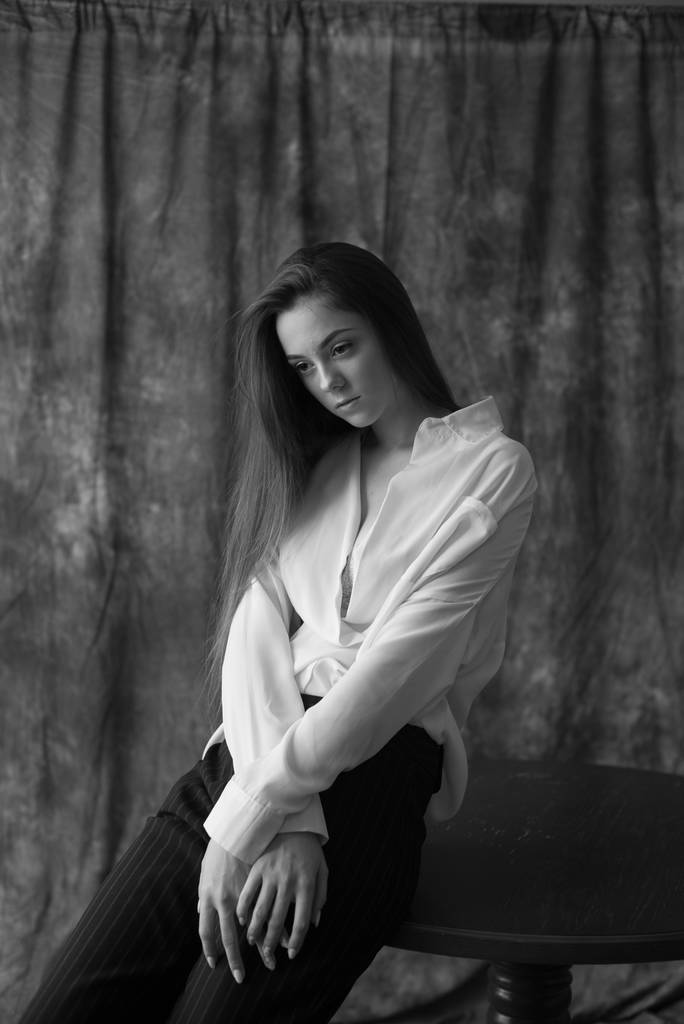 Υπέροχο μοντέλο νεαρής κοπέλας με σκούρα μακριά μαλλιά σε λευκό πουκάμισο και μαύρο παντελόνι που ποζάρει στο στούντιο σε υφασμάτινο φόντο. Ομορφιά και μόδα. - Φωτογραφία, εικόνα