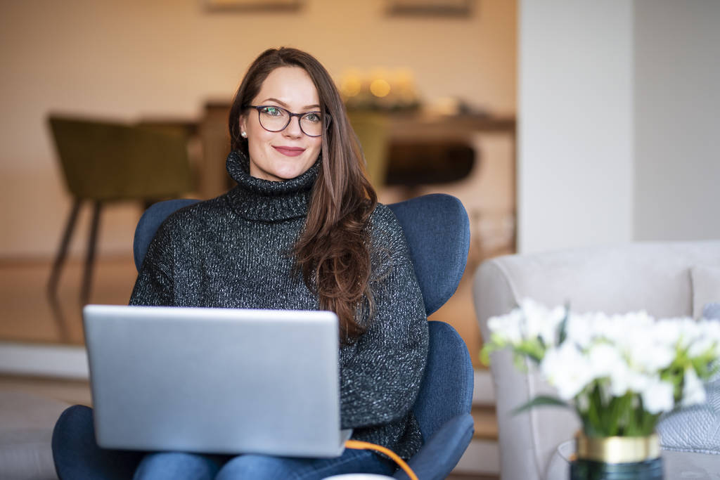Zdjęcie uśmiechniętej młodej kobiety korzystającej z laptopa podczas siedzenia na krześle i pracy w domu. - Zdjęcie, obraz