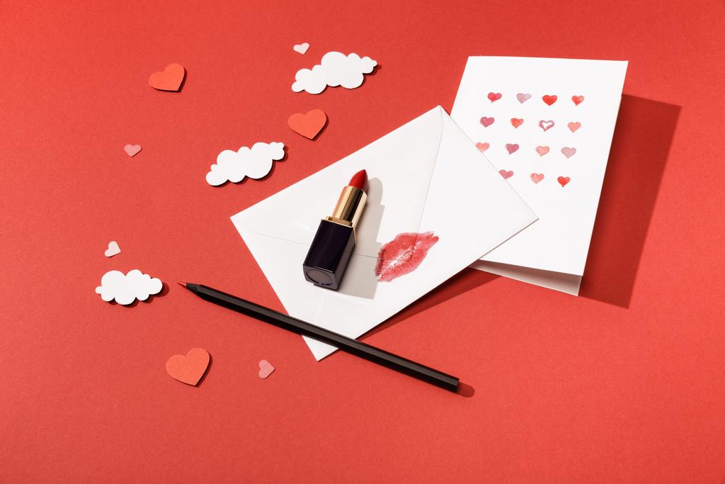 χάρτινα σύννεφα και καρδιές, ευχετήρια κάρτα κοντά σε φάκελο με σχέδιο χειλιών, κραγιόν και μολύβι σε κόκκινο φόντο - Φωτογραφία, εικόνα